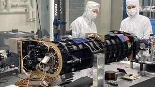 Two scientists in white coveralls examine a black piece of space tech with wires on one end.