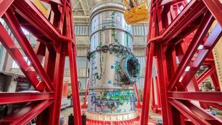 The cylindrical HALO module is seen standing vertically, surrounded by a red steel frame. The HALO module is silver and is lined with coils of green wires. 